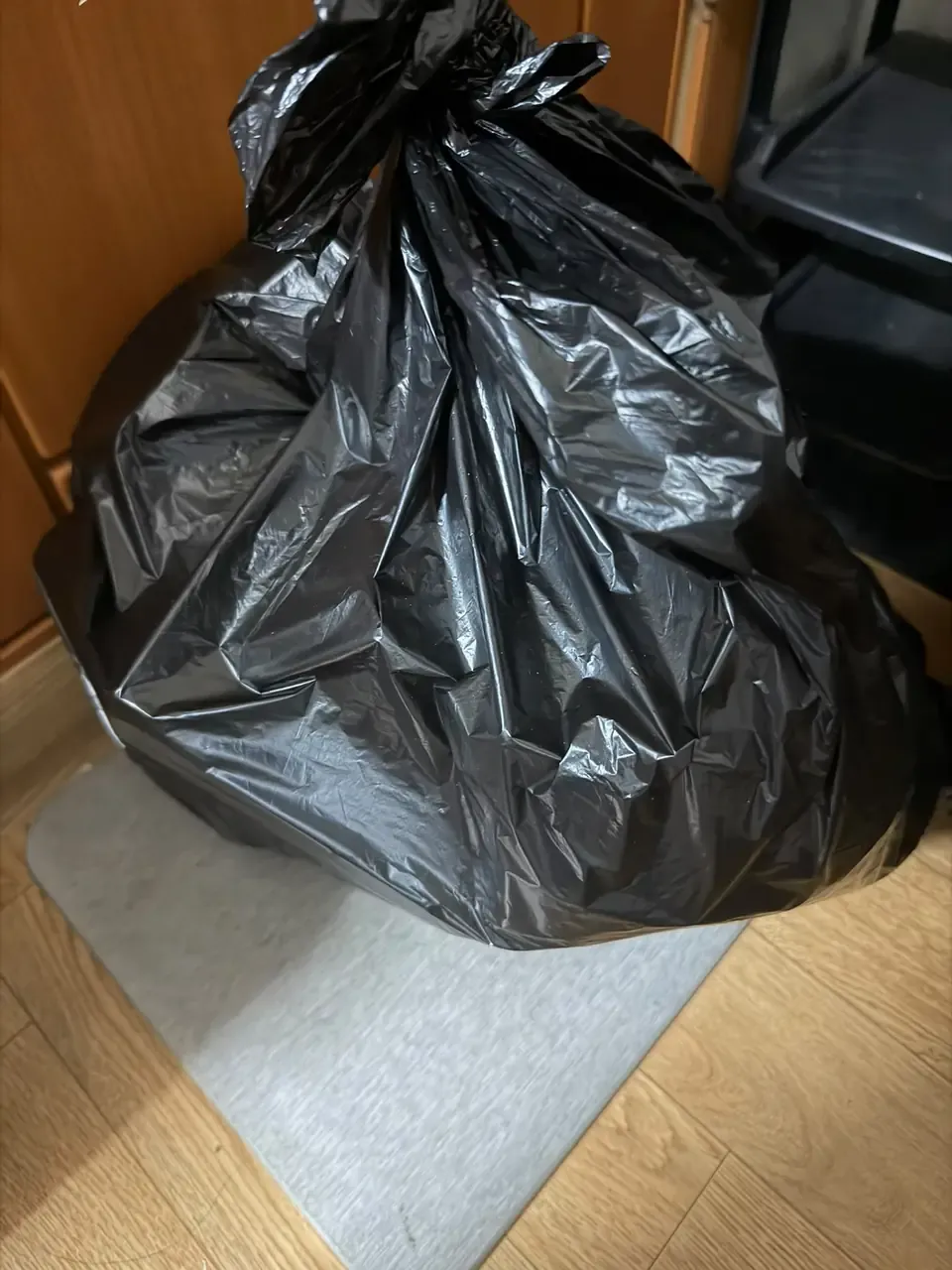 A tied black bag standing beside a shelf unit on the floor of a one-room