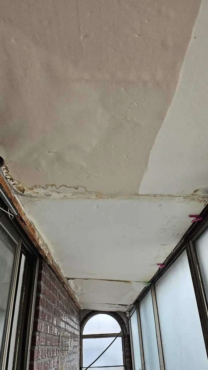 A balcony ceiling where the wallpaper has come away in a sheet, with brown water staining along the beam and rust on the window frame