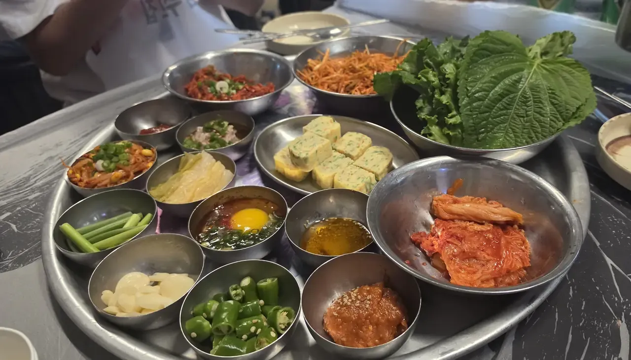 A steel tray of Korean side dishes including a bowl of whole-leaf cabbage kimchi, egg roll, garlic, green chillies, lettuce and perilla leaves