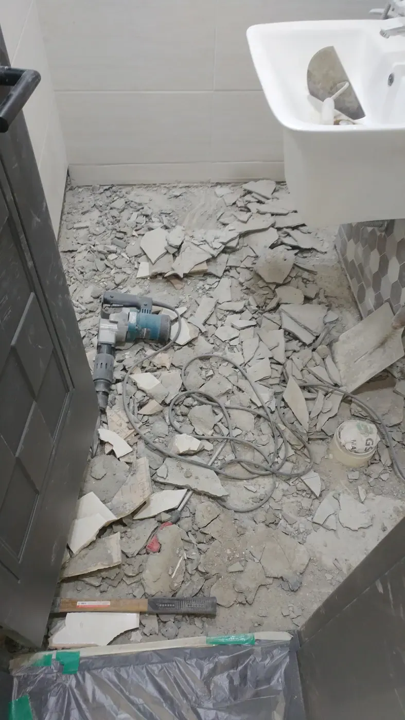 A Korean bathroom stripped back — tiles smashed, rubble across the floor, a demolition hammer lying where the floor used to be