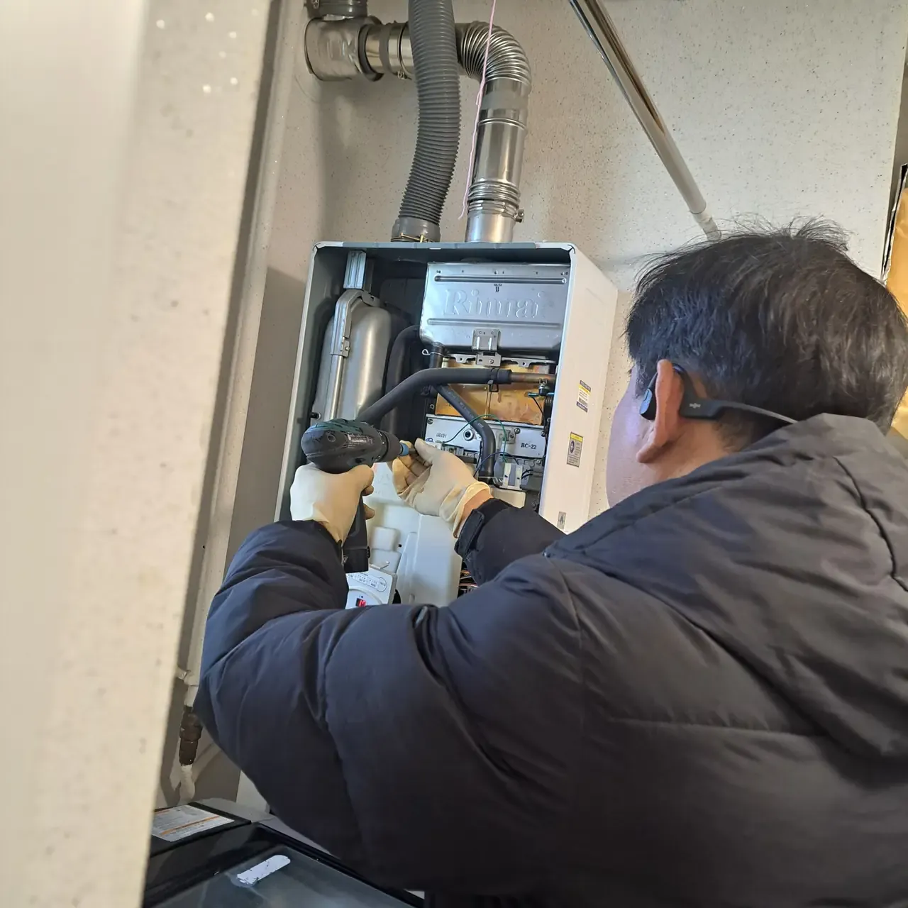 A technician kneeling at an open wall-mounted Rinnai gas boiler, working inside it with a cordless drill
