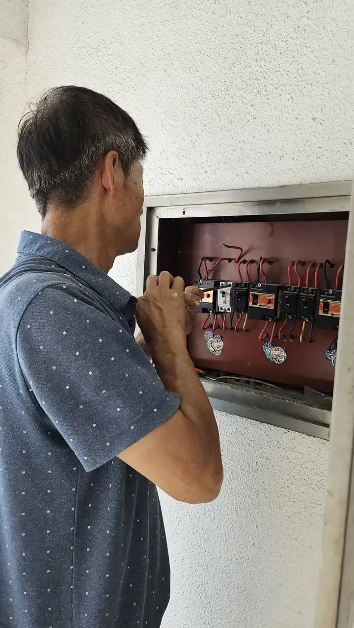 A man working inside an open breaker panel on an exterior wall, circuit breakers and wiring visible