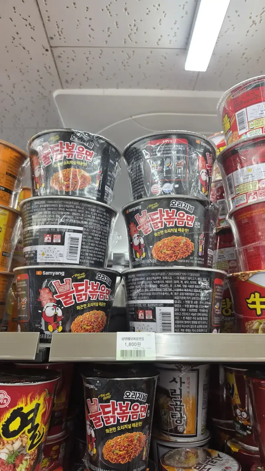 Stacked cups of buldak ramyeon on a convenience store shelf with a price tag reading 1,800 won