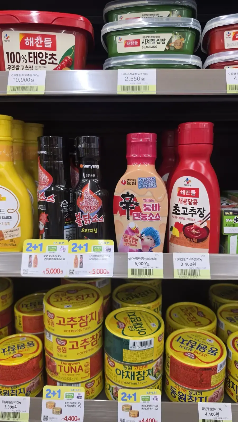 A convenience store condiment shelf with buldak sauce bottles, honey mustard and gochujang tubs, marked with 2+1 promotion tags