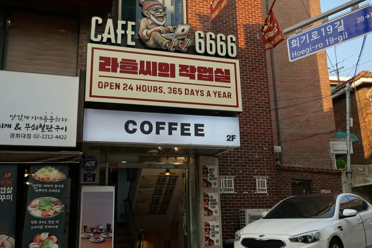 A cafe storefront near a Seoul university with a sign reading OPEN 24 HOURS, 365 DAYS A YEAR and a cartoon of a man working on a laptop