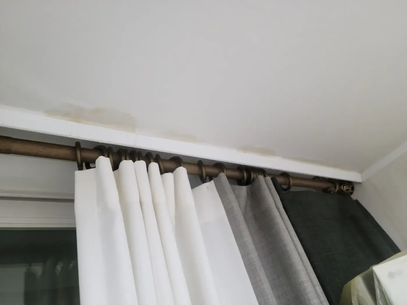 A pale brown water stain spreading along the ceiling edge above a curtain rail