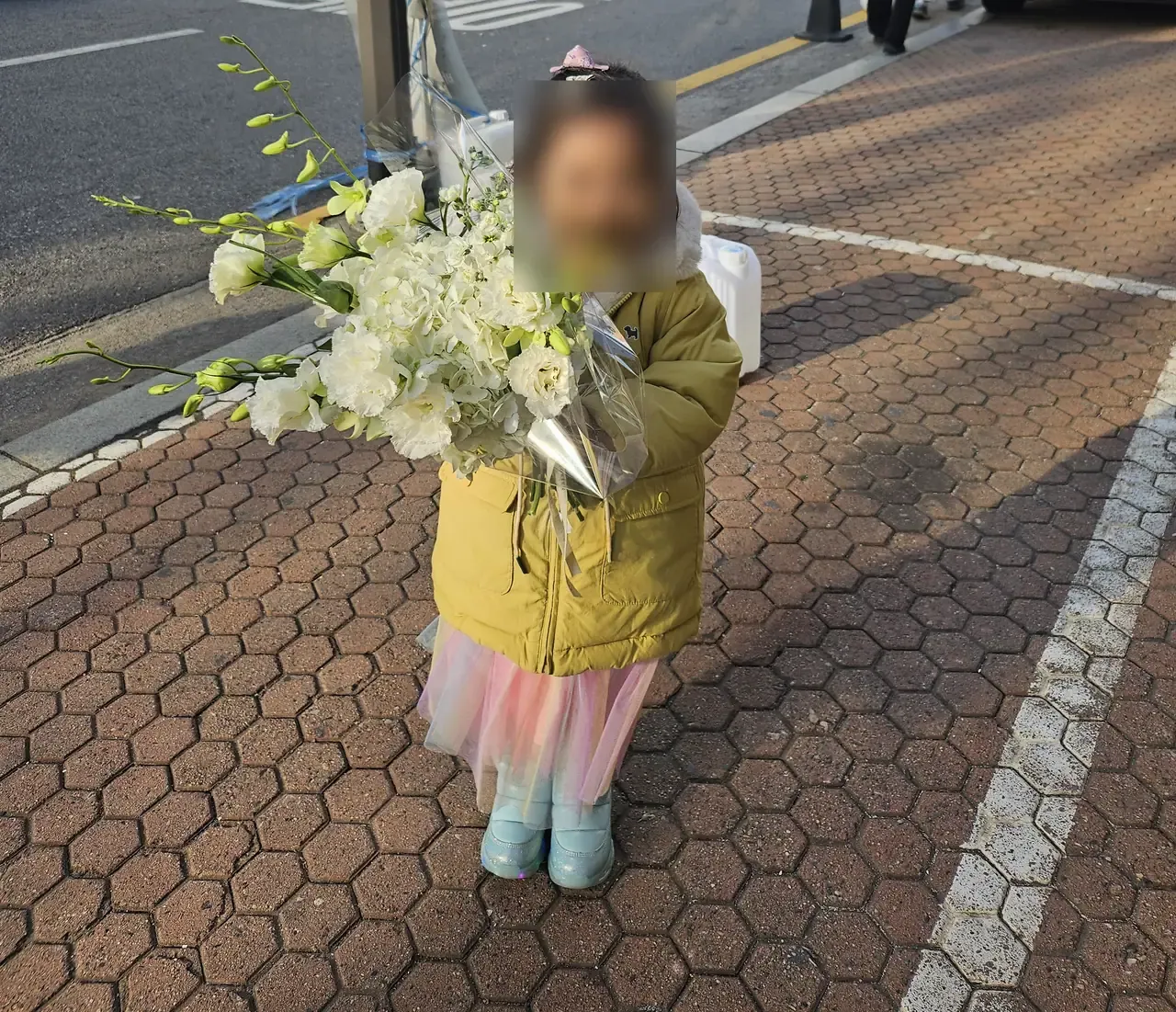 A young child in a padded coat standing on a Seoul pavement holding a large white bouquet