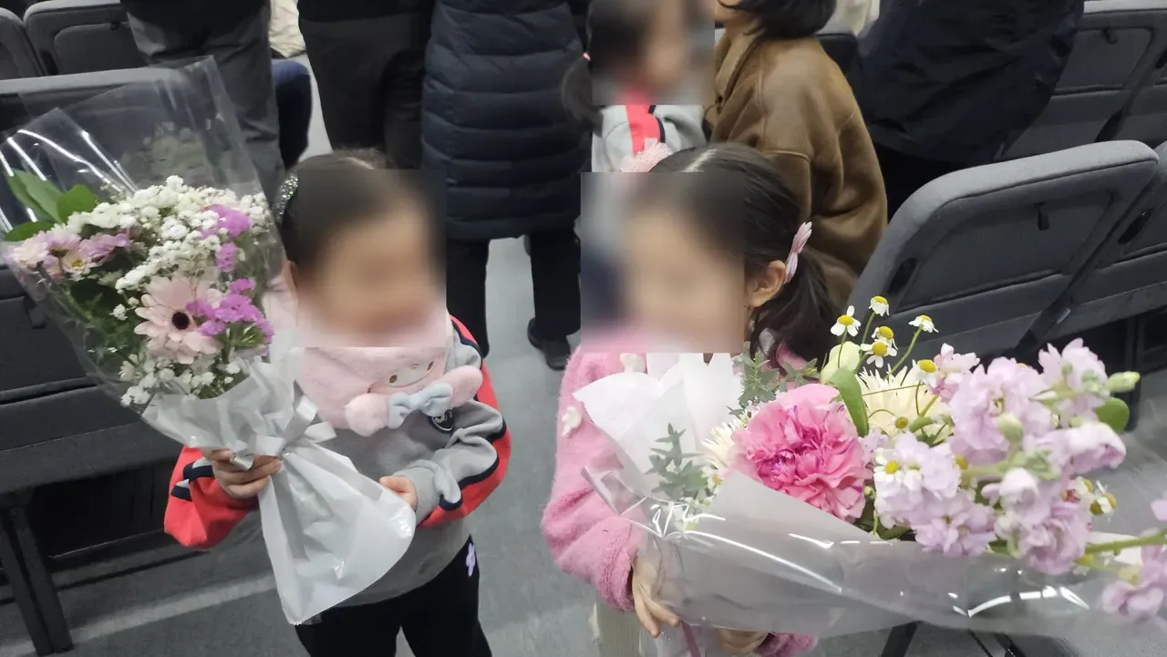 Two young children in winter coats holding wrapped bouquets at a ceremony, other families behind them