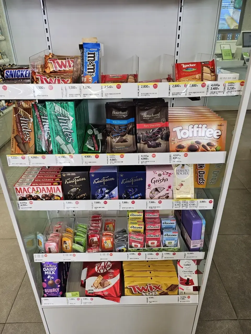 A convenience store chocolate shelf with red 1+1 and 2+1 tags beside the prices and a yellow sale tag showing a reduced price