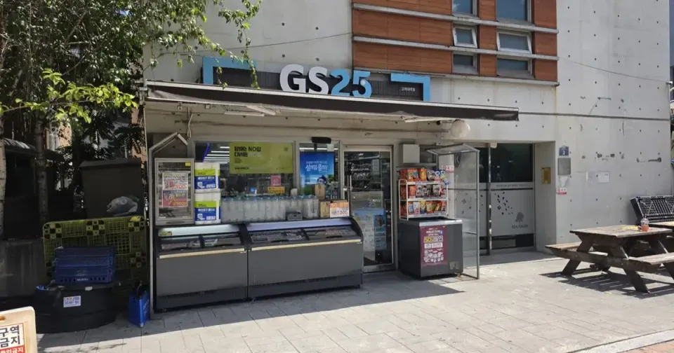 A GS25 convenience store on a Seoul street in daylight, with an outdoor bench and tables in front