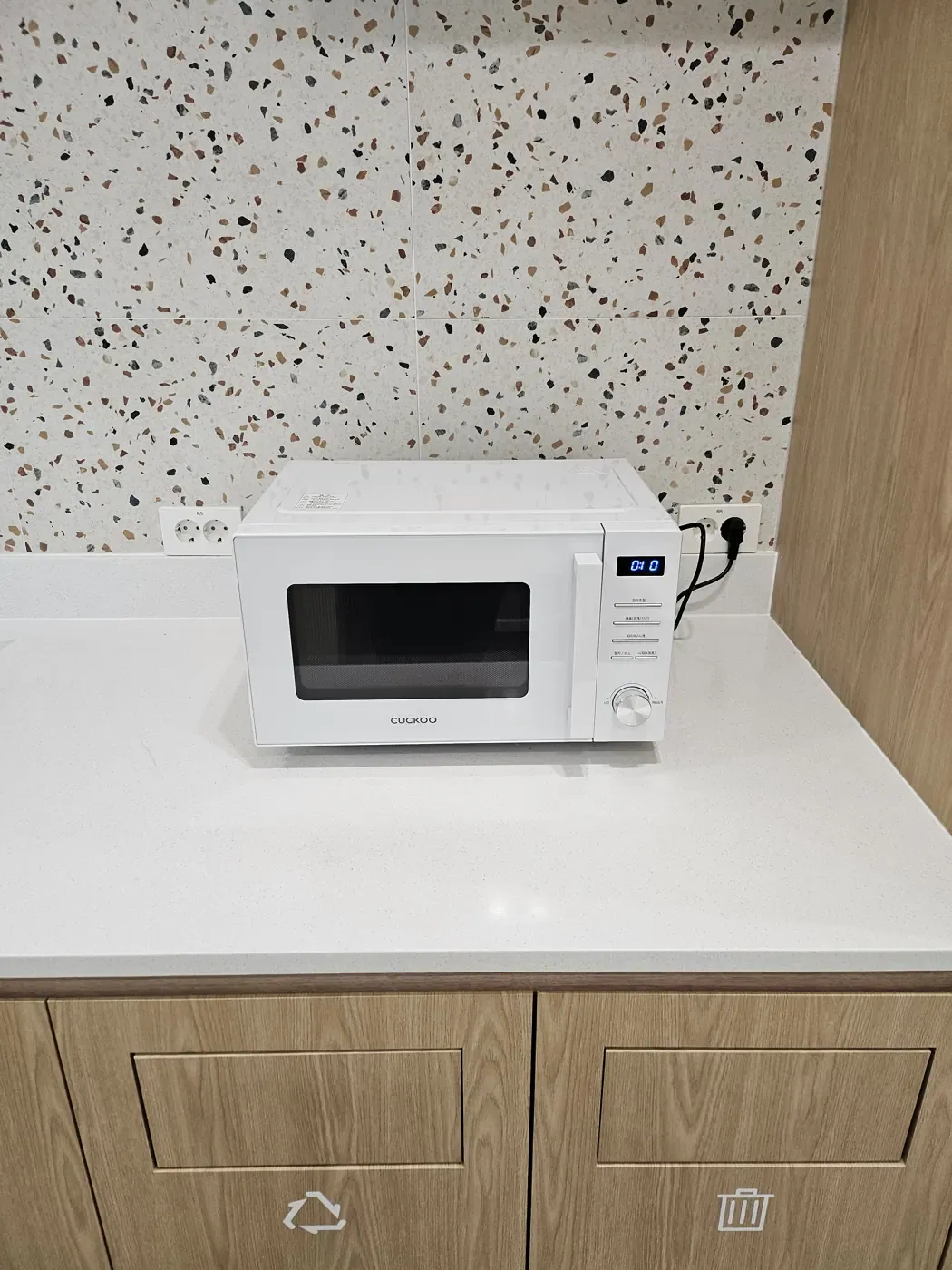 A microwave on a convenience store counter, above cabinet doors marked with recycling and waste bin symbols