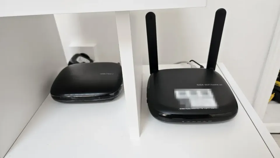 A KT GiGA WiFi home ax router beside its fibre terminal in a cupboard, the credential label deliberately blurred