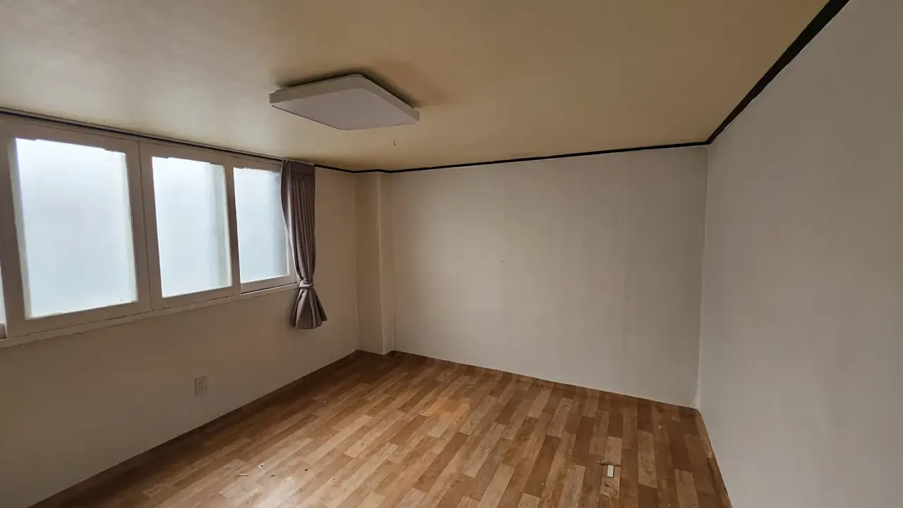 An empty Korean room at handover with frosted windows, a curtain left behind, a square ceiling light and wood-effect flooring with grit scattered across it