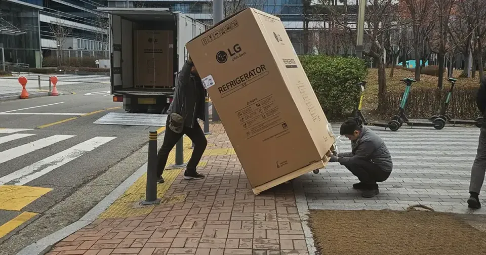 Two movers easing a boxed LG refrigerator off a truck onto a Seoul pavement