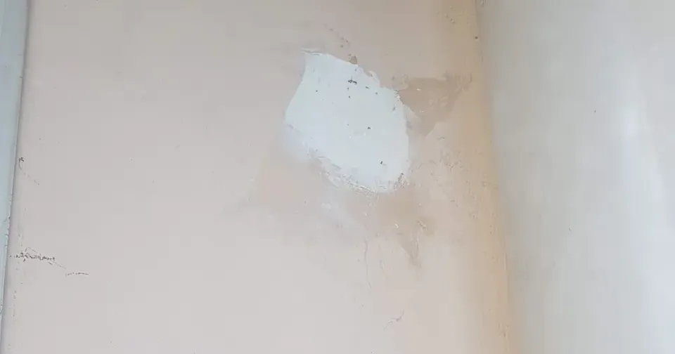 A beige wall with a rough white putty patch where a hole was filled in, clearly visible against the surrounding wallpaper