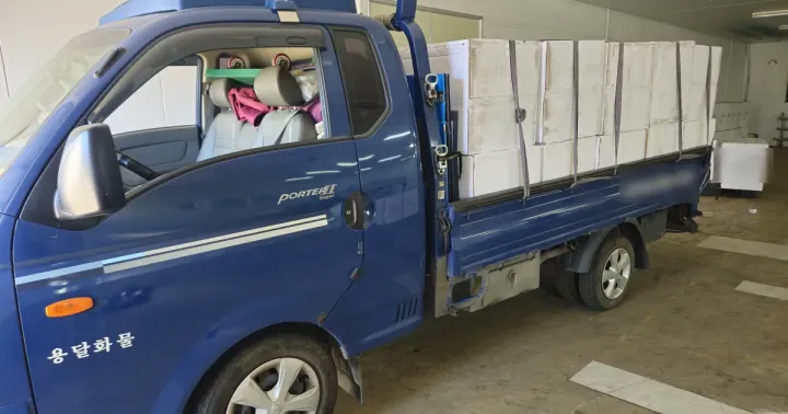 A blue Hyundai Porter 1-ton truck with its bed loaded to the top rail with white cardboard boxes, held by black straps