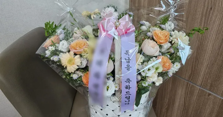 A white basket arrangement of roses, gerberas and lisianthus with two vertical purple ribbons, the right one reading in Korean: congratulations on the birth of your son