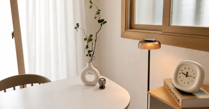 A corner of the shared common room: a dining table, a vase, a lit floor lamp and a clock on the sideboard beside a curtained window