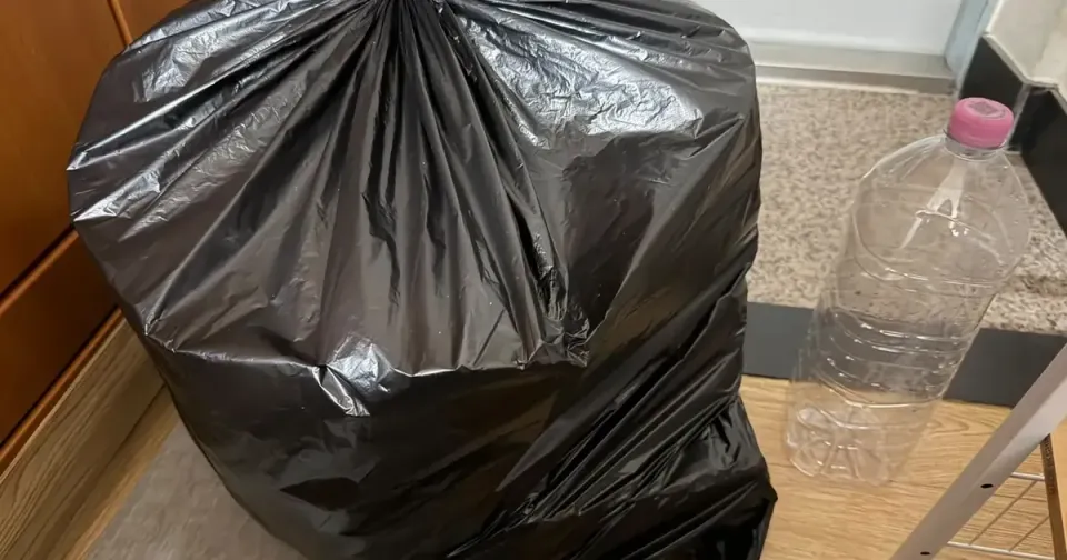 A tied black rubbish bag on the floor of a small Korean flat, beside a cabinet and a plastic bottle