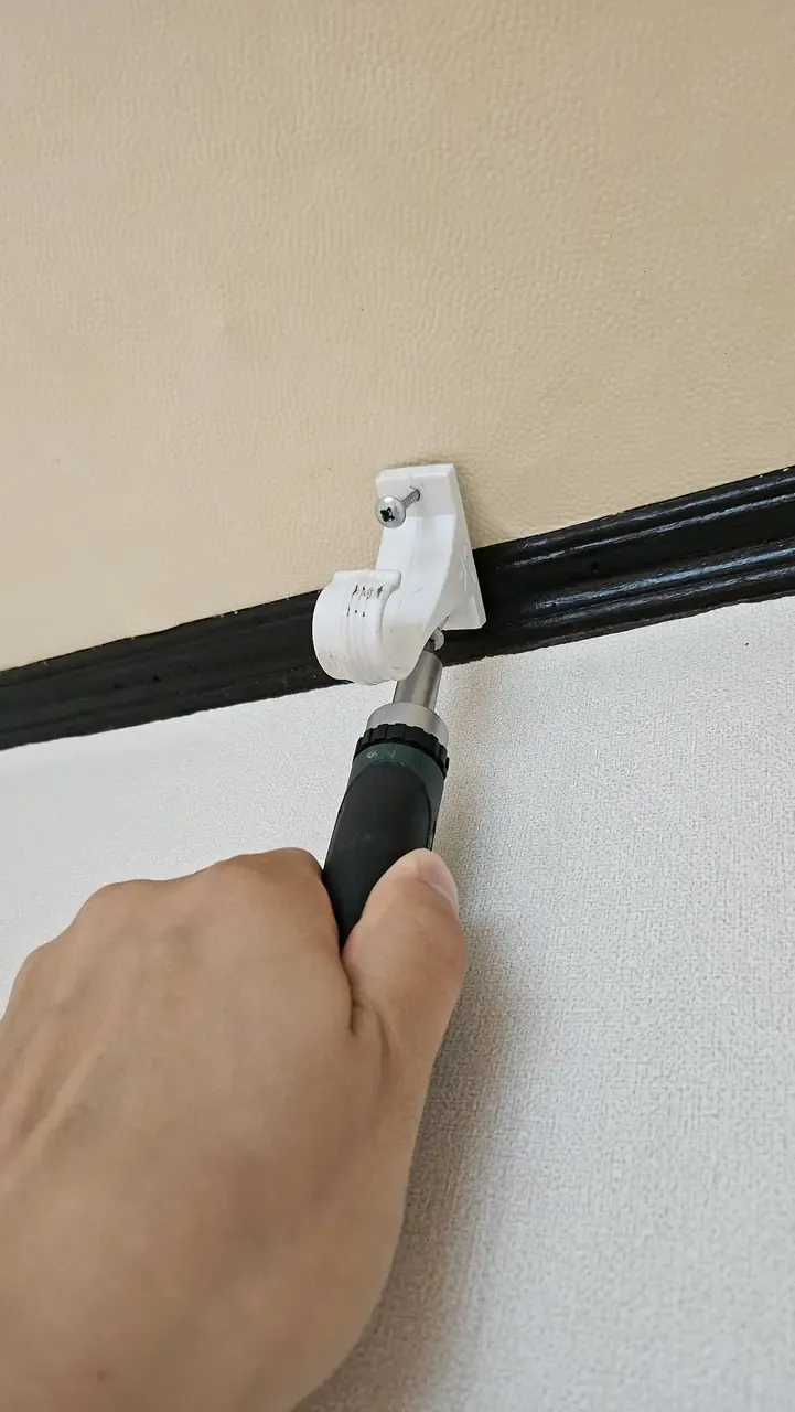 A hand driving a screw into a white curtain-rail bracket at the top of a wall