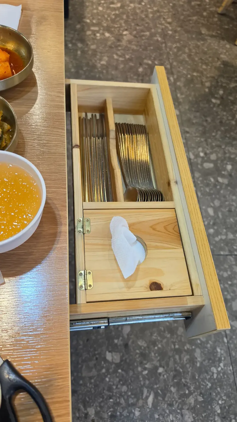 An open cutlery drawer pulled out from the side of a Korean restaurant table, holding chopsticks, spoons and a napkin dispenser