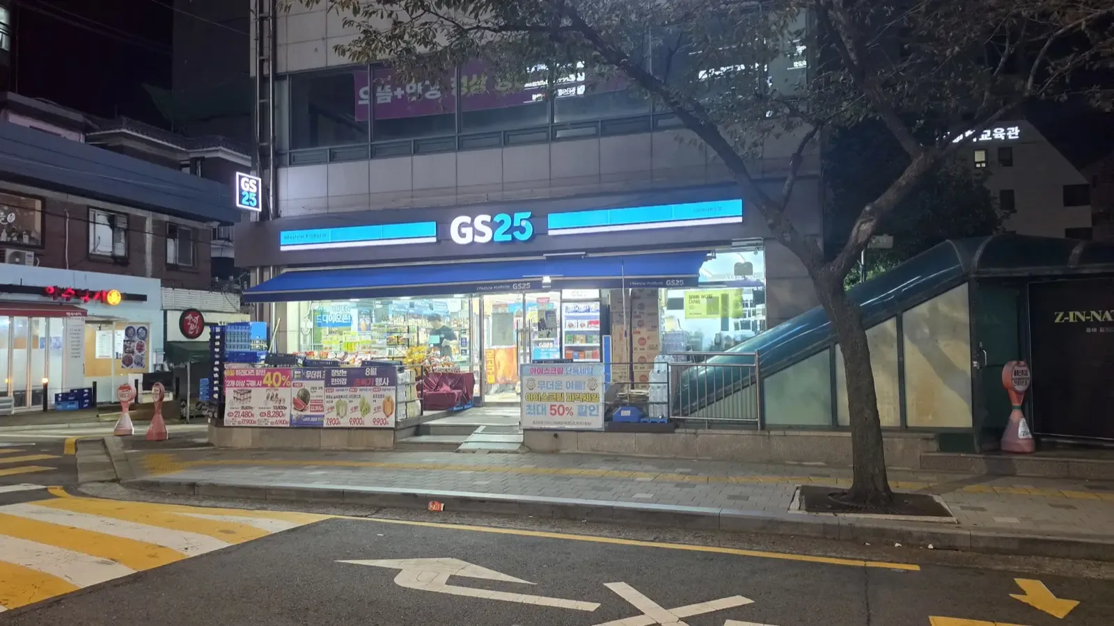 A GS25 convenience store lit up on an empty street late at night, with a subway entrance beside it