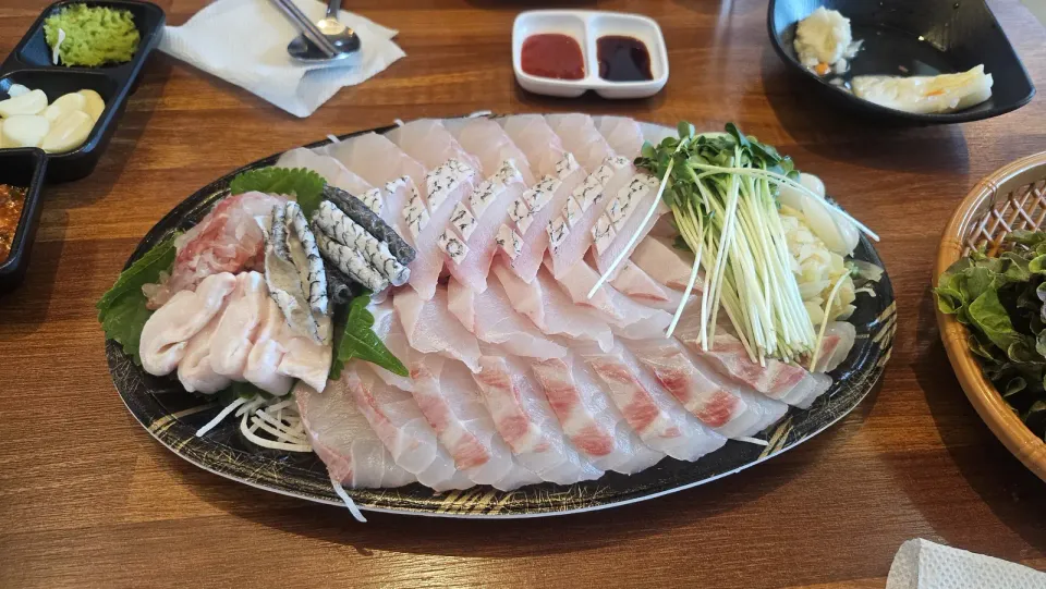 A large oval platter of thinly sliced white raw fish with perilla leaves, radish and dipping sauces on a wooden table in Seoul