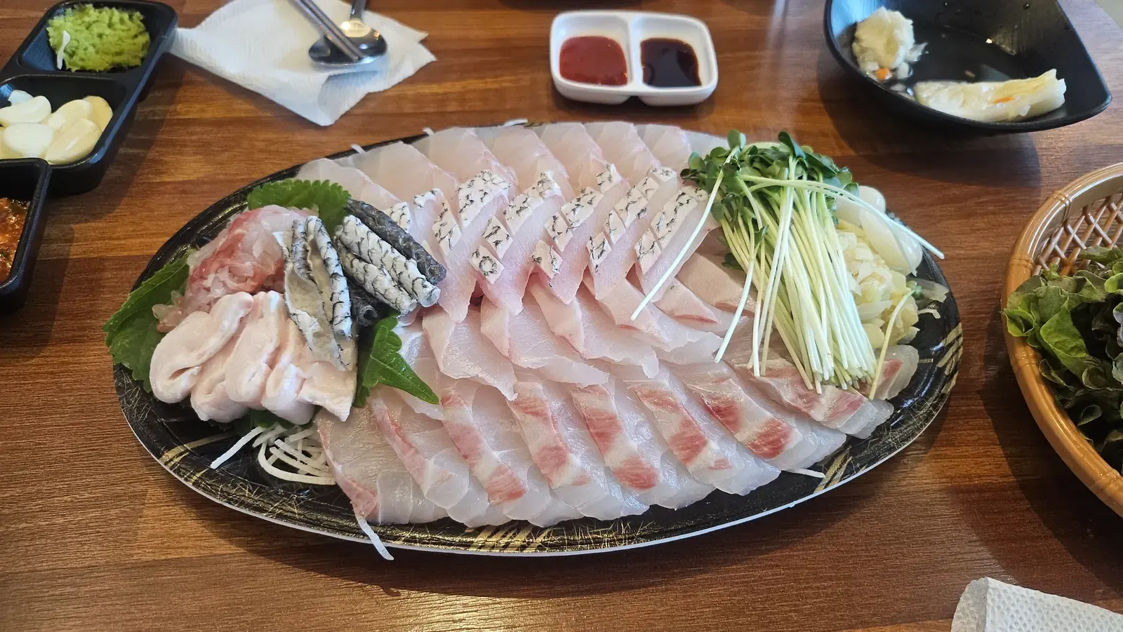 A large oval platter of sliced white raw fish with perilla leaves, sprouts, radish and two dipping sauces
