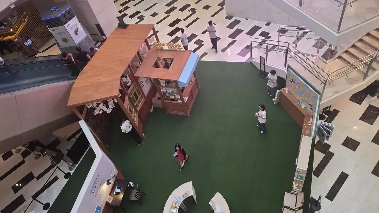 Overhead view of a wooden pop-up bookshop on green turf in the IFC Mall Yeouido atrium, with reading benches and a sticker wall