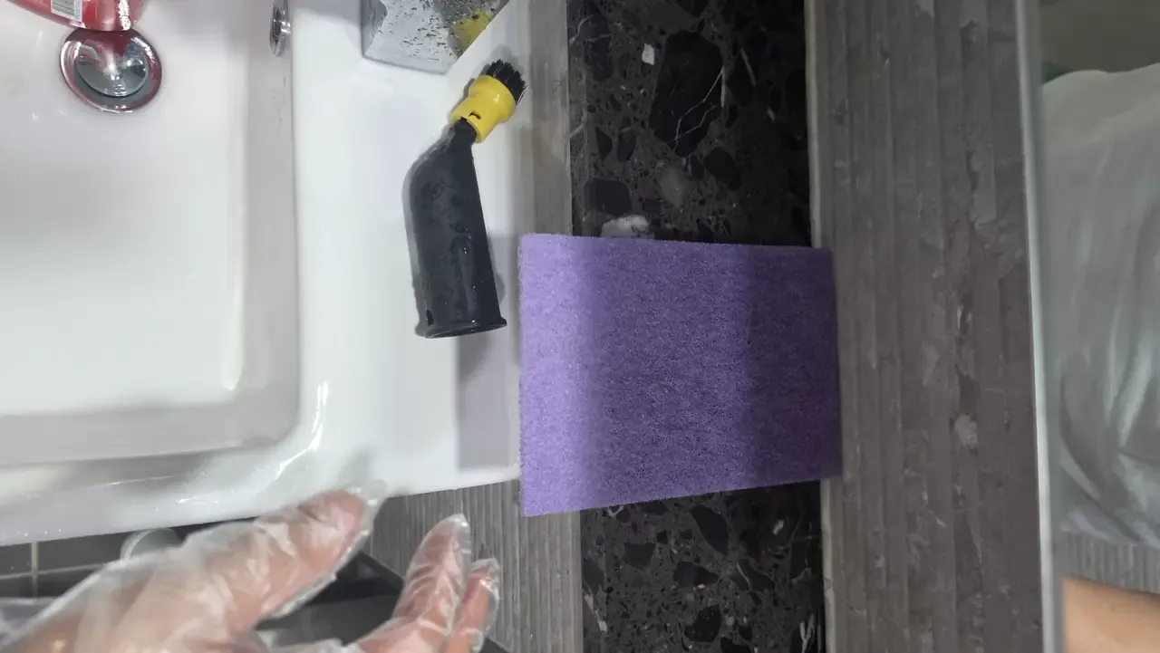 A steam cleaner brush head and a purple scouring pad on a basin edge