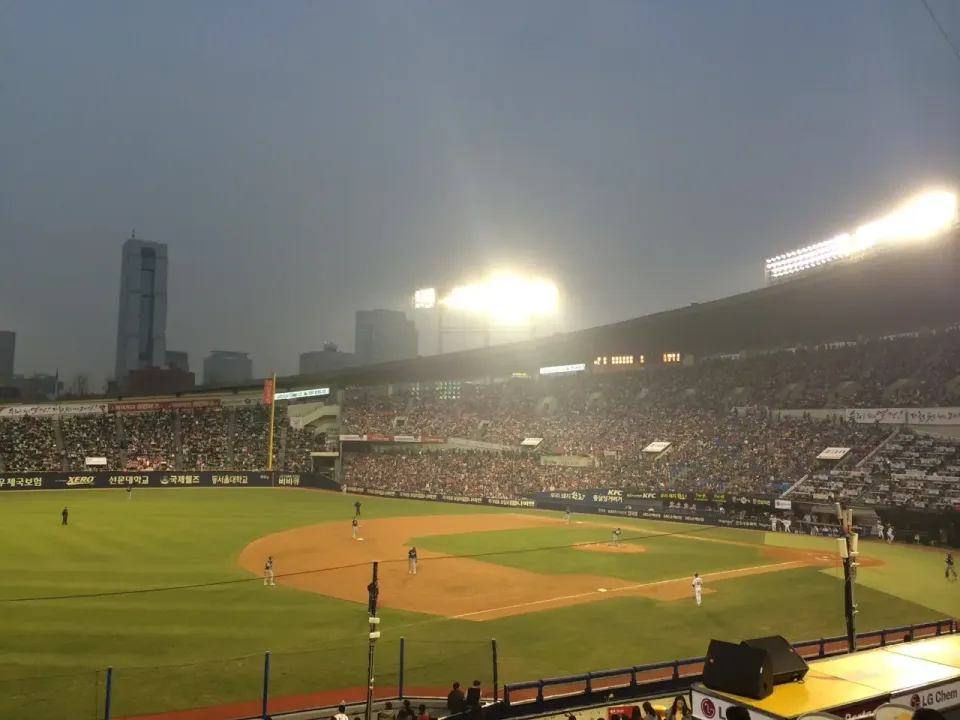 Packed KBO baseball stadium in Seoul under floodlights at dusk, with the city skyline behind the stands