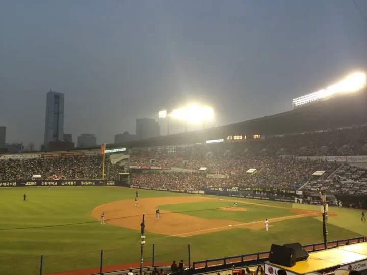 Packed KBO baseball stadium in Seoul under floodlights at dusk, with the city skyline behind the stands