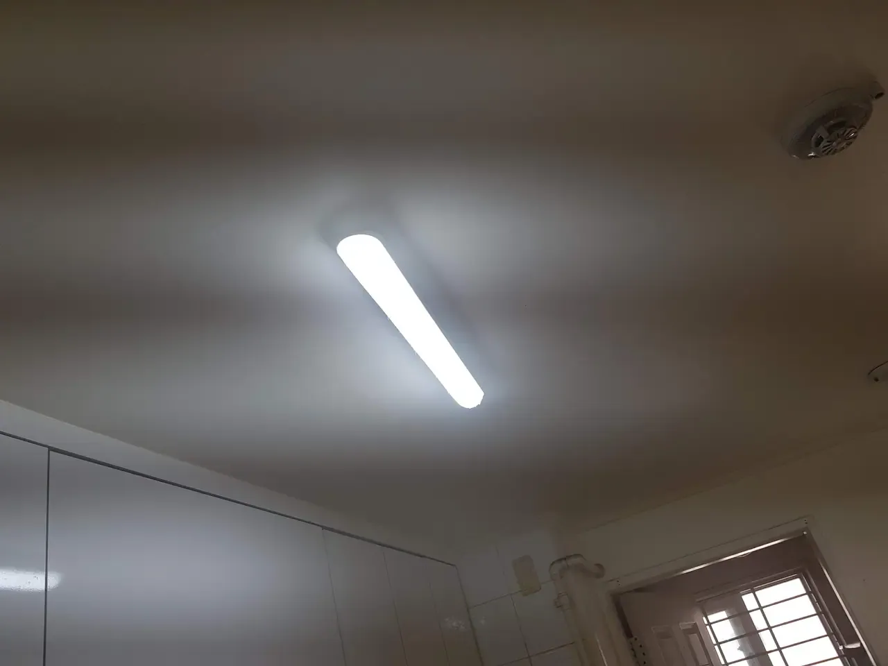 A white kitchen ceiling with a long LED light fitting and a smoke detector, cabinet tops visible below