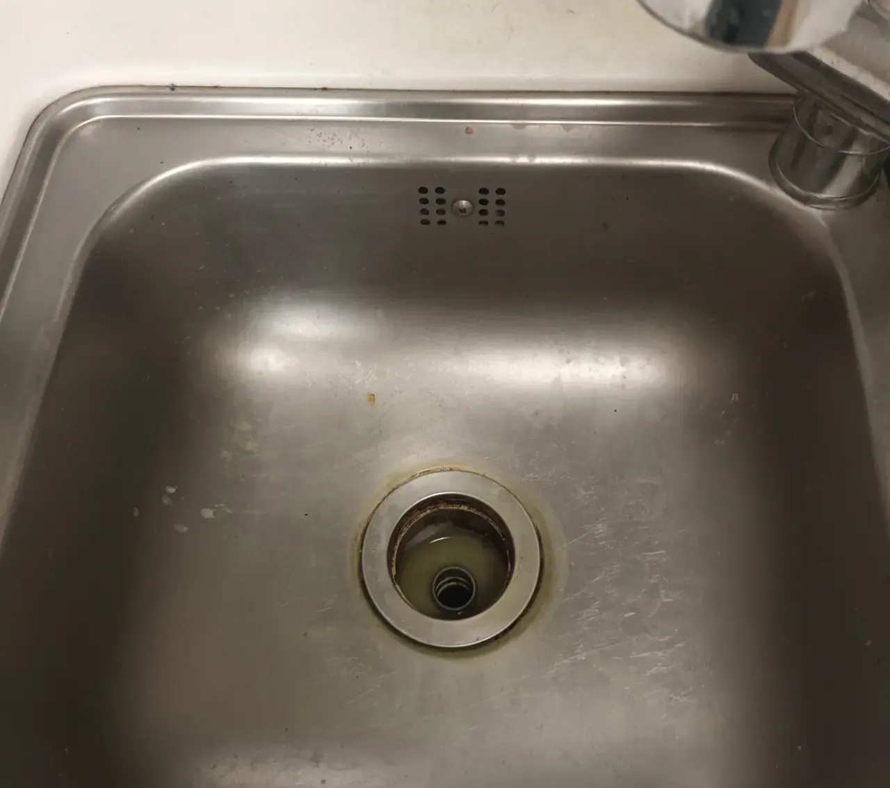 A stainless steel Korean kitchen sink with the drain strainer lifted out, showing brown grease build-up around the drain opening