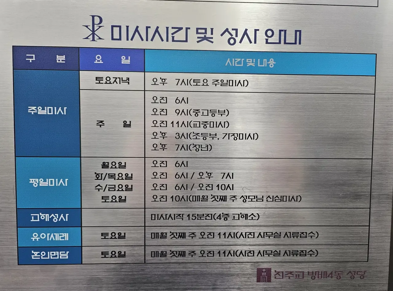 Stainless-steel Mass timetable board outside a Seoul parish church, listing Sunday, weekday, confession and baptism times in Korean