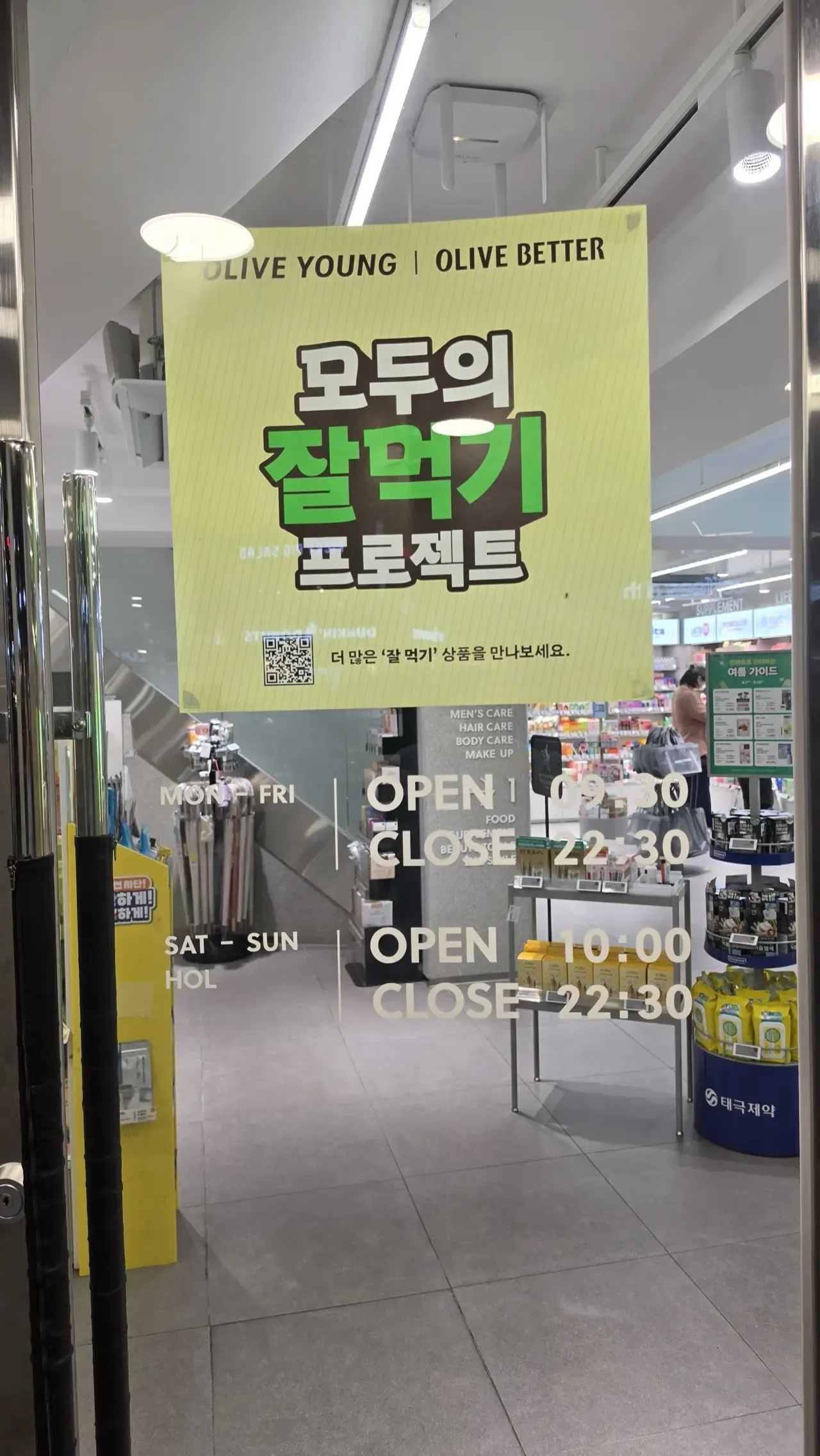 The opening-hours sign on an Olive Young door reading Mon–Fri open 09:30 close 22:30, Sat–Sun and holidays open 10:00 close 22:30