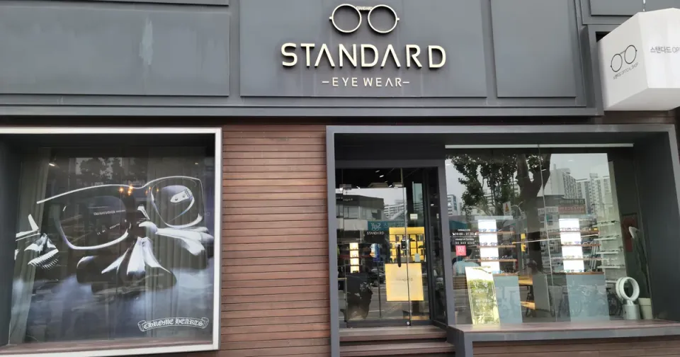 The Standard Eyewear optical shop at street level near Isu Station in Seoul, its window showing a wall of frames and opening hours of 10am to 9pm