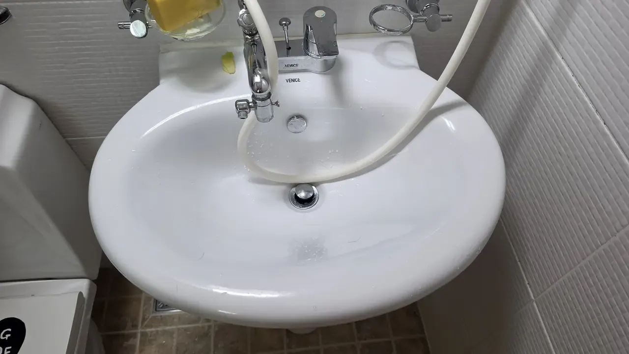 A white pedestal basin in a Korean bathroom with a chrome pop-up stopper set into the drain and a bidet hose running across it