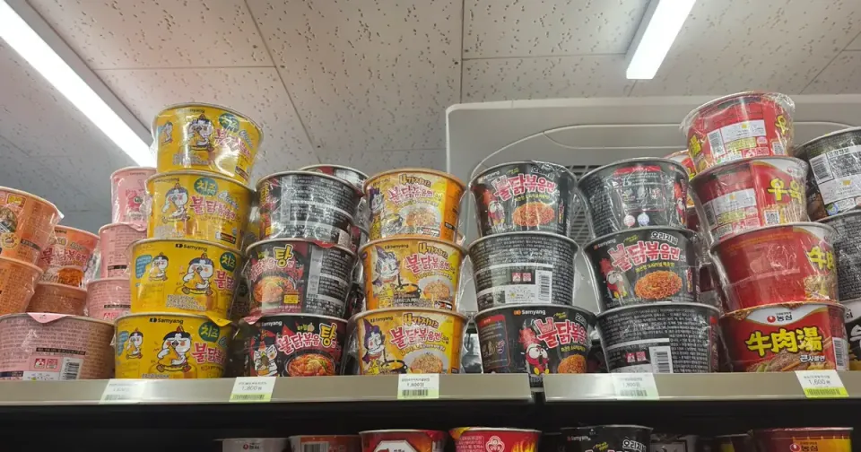 A convenience store shelf stacked with Korean cup ramyeon — cheese buldak, carbonara buldak, mala buldak, original buldak and beef soup cups, priced at 1,800 won