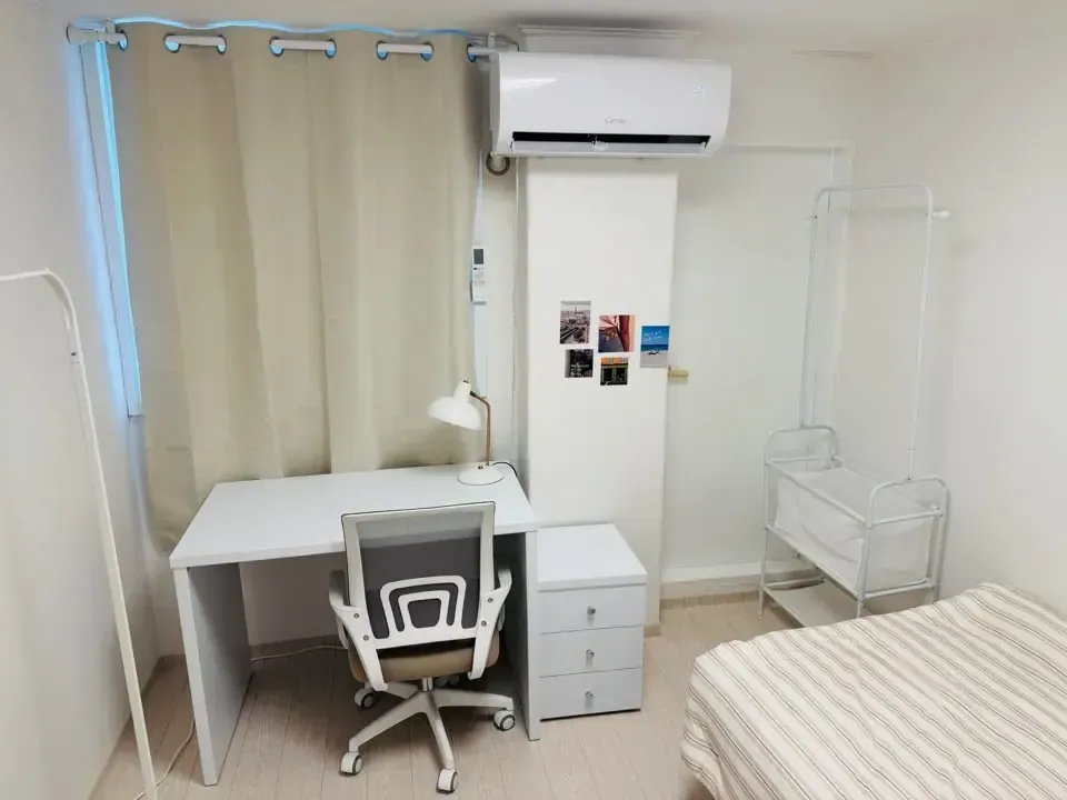 A room with a wall air conditioner, a desk and chair, and a freestanding clothes rail beside the bed