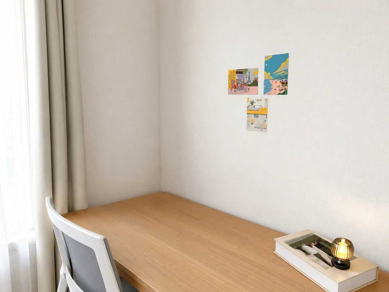 A desk and chair by the window, with a few postcards on the wall above it