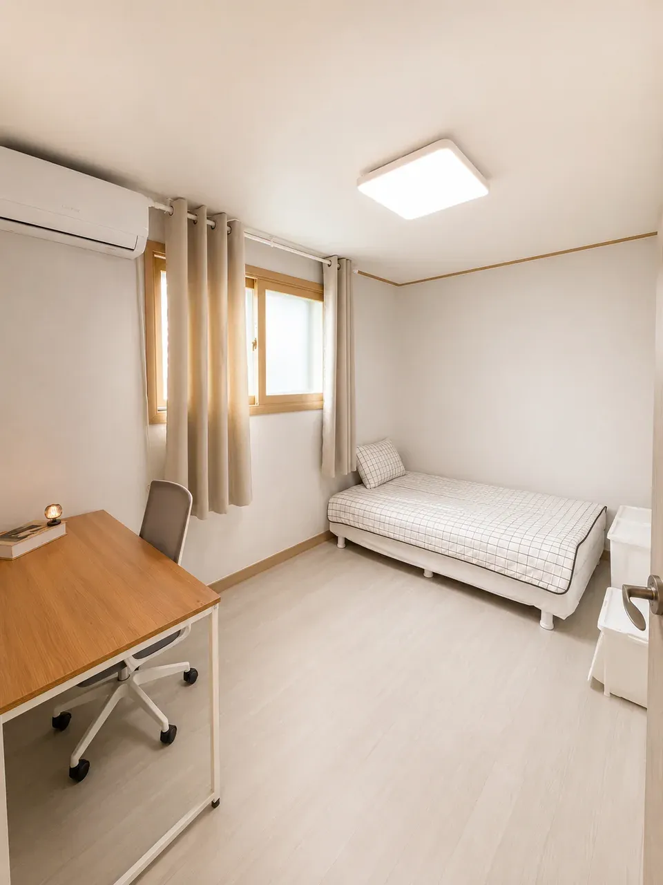 A bright single room with the desk in the foreground by the door, the bed against the far wall and a wall air conditioner above