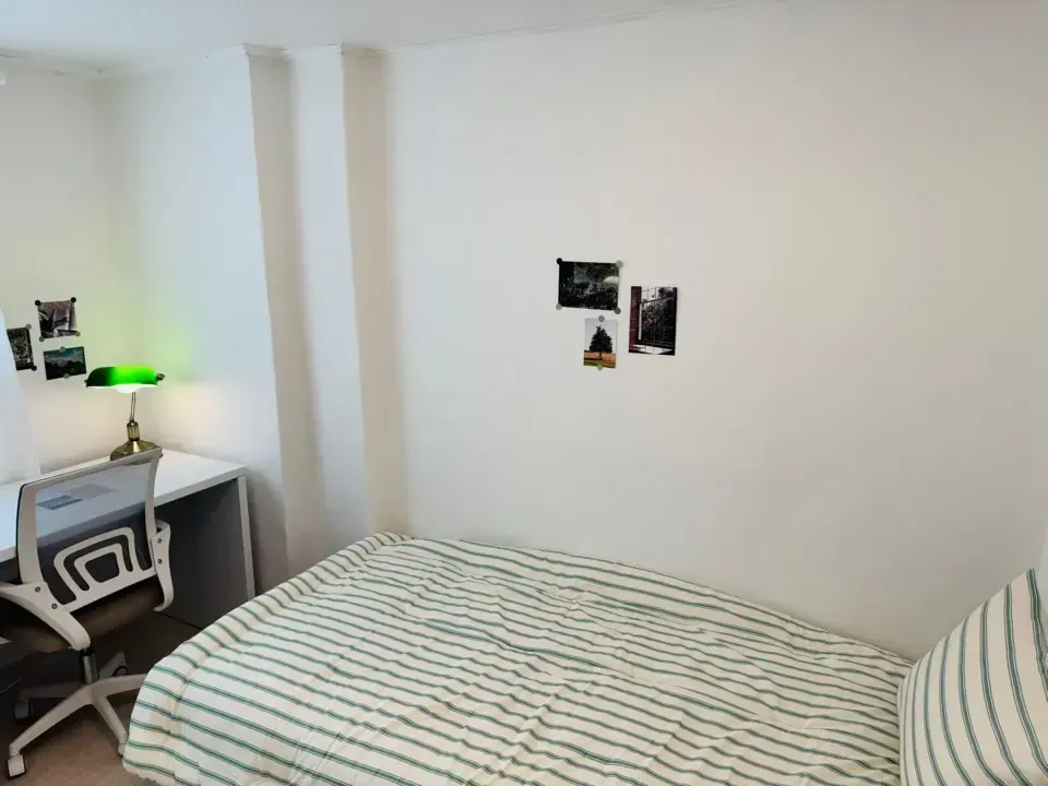 A single room with a bed, a desk with a green lamp and postcards on the wall