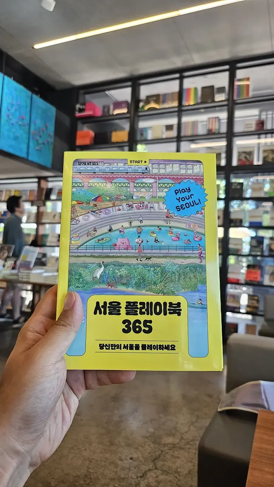 A hand holding a yellow booklet titled Seoul Play Book 365 — Play Your Seoul, inside a book cafe