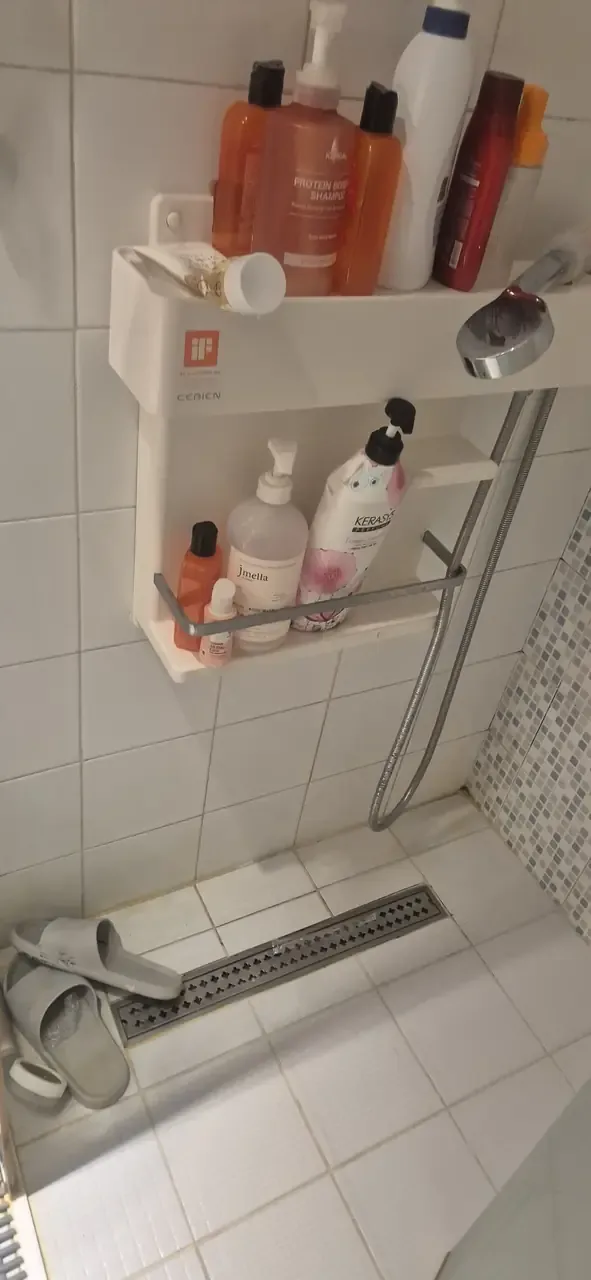 A shower shelf loaded with bottles above a tiled floor and linear drain, before cleaning