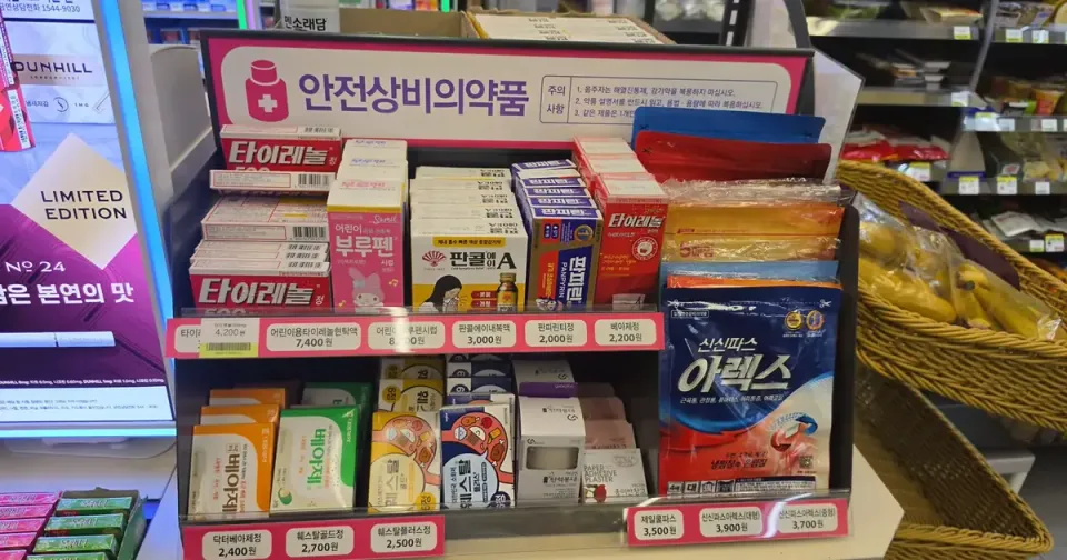 A convenience store stand labelled 안전상비의약품 holding Tylenol, children's fever syrups, cold remedies, digestive tablets and pain patches with price tags