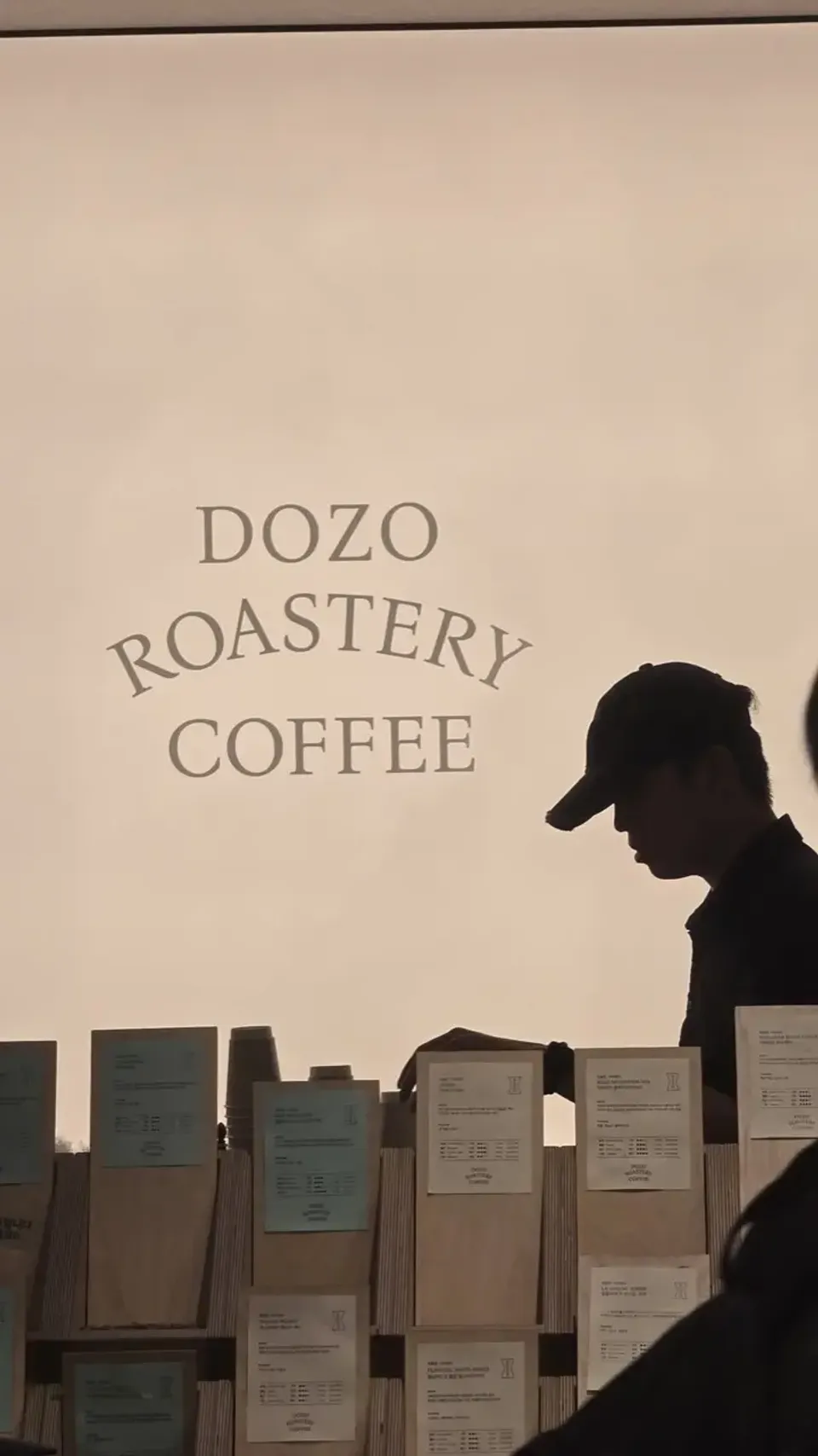 A barista silhouetted against a lit wall at a Korean specialty coffee roastery, with bags of beans on shelves