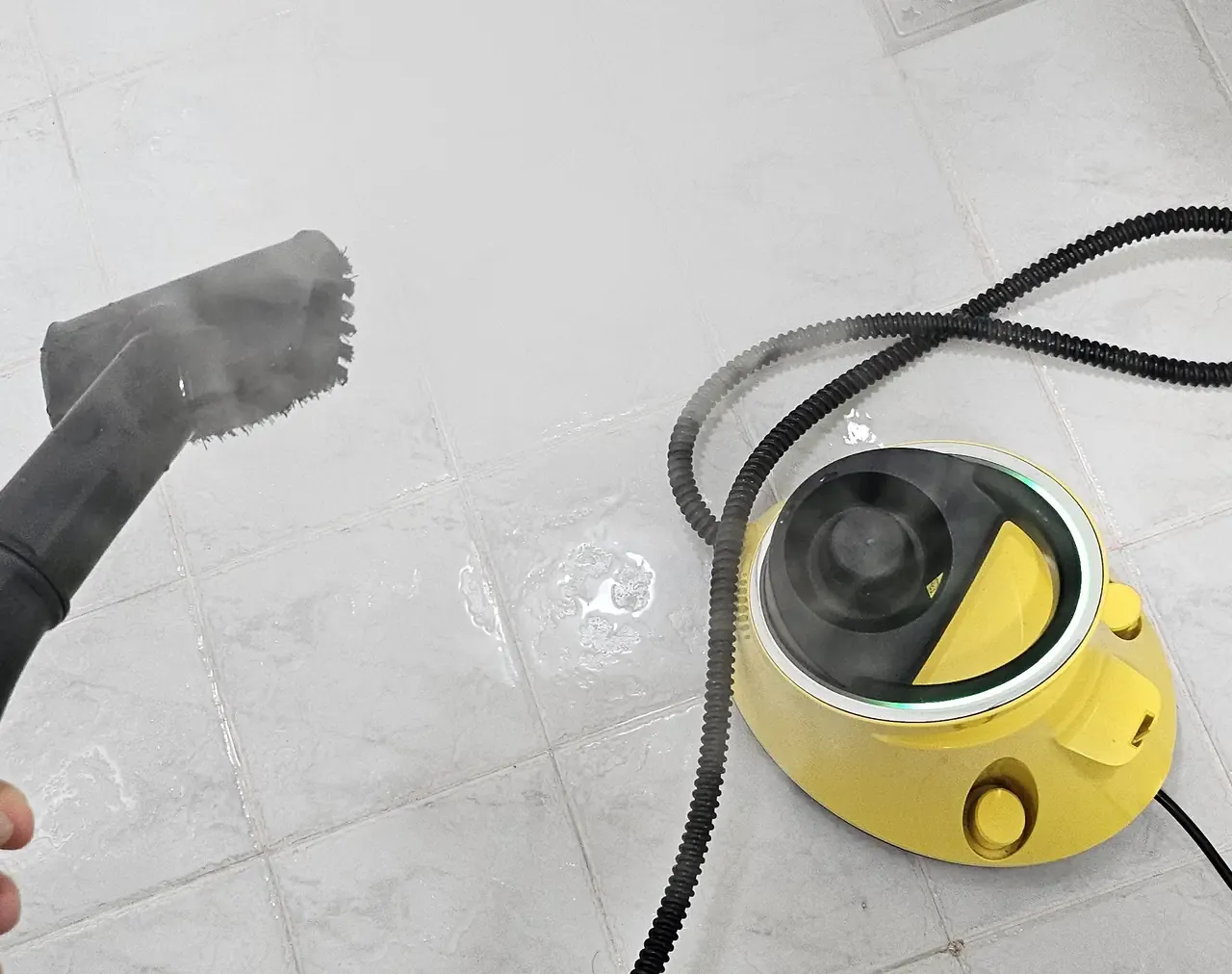 A handheld steam cleaner nozzle releasing steam over a tiled bathroom floor, the yellow domestic steam unit and its coiled hose beside it