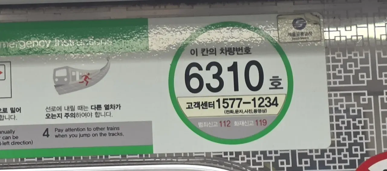 A green circular sticker inside a Seoul subway carriage reading 이 칸의 차량번호 6310호, next to the emergency instructions panel