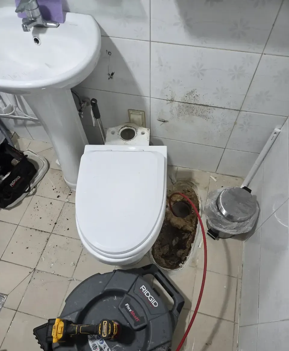 A Korean bathroom with the toilet lifted off the floor and stood to one side, the open waste flange thick with build-up, a drain machine and tool bag on the tiles